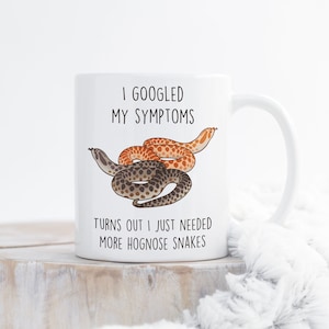May include: White ceramic mug featuring the text "I Googled My Symptoms Turns Out I Just Needed More Hognose Snakes." The mug has a cartoon illustration of two hognose snakes, one brown and one orange. The mug has a white handle.