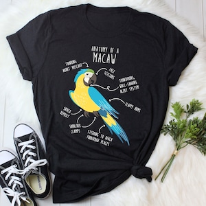 Puede incluir: Una camiseta negra con un gráfico de un guacamayo azul y amarillo. El gráfico incluye texto que dice "Anatomía de un Guacamayo" y etiqueta diferentes partes del ave, como "Tijeras de la cara", "Sistema de alerta atronador, que sacude las paredes" y "Dirección para llegar a lugares prohibidos".