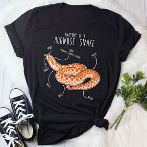Könnte beinhalten: Ein schwarzes T-Shirt mit einer weißen Grafik eines Hognose-Schlangens mit dem Text "Anatomie einer Hognose-Schlange" und einem Diagramm der Körperteile der Schlange, die mit "Smoot", "Read (Prammatic Troikats Inside)", "PBBBT", "Neck" und "Neck?" beschriftet sind.