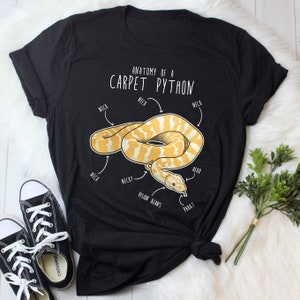 Puede incluir: Una camiseta negra con un gráfico blanco de una pitón alfombra amarilla y blanca con el texto "Anatomy of a Carpet Python" y etiquetas para "Neck", "Neck?", "Vision Beans", "Read" y "PBBBT".