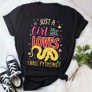 May include: Black t-shirt with the text "Just a girl who loves ball pythons" in pink, red, and yellow lettering. A yellow ball python is illustrated on the shirt.