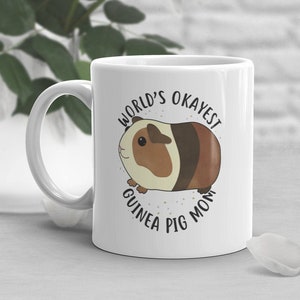 May include: White ceramic mug with a brown and white guinea pig illustration and the text "World's Okayest Guinea Pig Mom".