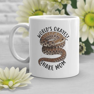 Pode incluir: Caneca de café de cerâmica branca com uma ilustração de cobra preta e marrom e o texto "World's Okayest Snake Mom".
