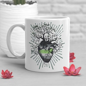 May include: White ceramic mug featuring a stylized illustration of a heart with a tree growing from it. The heart design incorporates the colors of the aromantic pride flag: green, gray, and black. The design is surrounded by green rays.