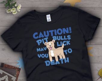 Op de afbeelding: Zwart t-shirt met de tekst "CAUTION! PIT BULLS MAY LICK YOU TO DEATH" in blauw. Een cartoon pitbull met een roze tong staat in het midden. Het shirt is gemaakt van een zacht materiaal.