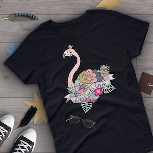Puede incluir: Camiseta negra con un flamenco rosa que lleva una corona y está rodeado de flores. El texto "Zero Flocks Given" está en una pancarta debajo del flamenco.