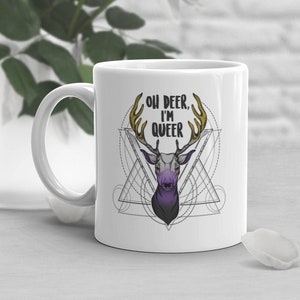 May include: White ceramic mug featuring a deer design and the words "OH DEER, I'M QUEER." The deer has gold antlers and a purple and grey face, set against a geometric pattern. The mug has a curved handle.