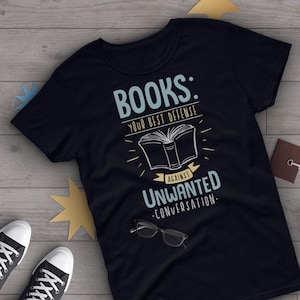 以下が含まれることがあります： 黒いTシャツに、白い文字で「本：不要な会話に対する最高の防御」と書かれています。グラフィックには、開いた本のスタイリッシュな画像が含まれています。
