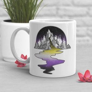 Puede incluir: Taza de cerámica blanca con una ilustración en blanco y negro de una cordillera reflejada en un cuerpo de agua. El agua está coloreada con los colores amarillo, blanco y morado de la bandera del orgullo no binario.