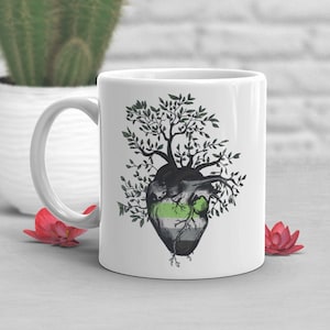 May include: White ceramic mug with a green, white, and black heart design. The heart is surrounded by a tree with black branches and green leaves.