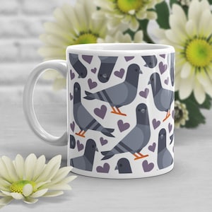 May include: White ceramic mug featuring a repeating pattern of gray pigeons and purple hearts. The mug has a white handle and is set against a background of white flowers and a white brick wall.