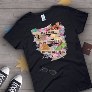 Op de afbeelding: Zwart t-shirt met een kleurrijke afbeelding van verschillende reptielen en de tekst "ALL I WANT IS TO CUDDLE ALL OF THE REPTILES". Het ontwerp bevat een slang, een hagedis, een schildpad en bloemen.