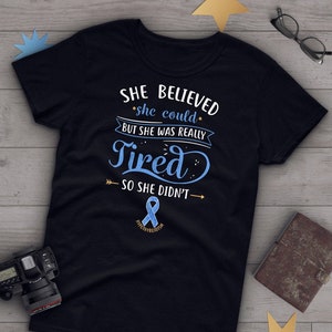 Puede incluir: Camiseta negra con una cinta azul y el texto "SHE BELIEVED she could BUT SHE WAS REALLY Tired SO SHE DIDN'T" con una flecha apuntando hacia la izquierda y una flecha apuntando hacia la derecha.