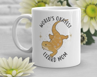 Puede incluir: Taza de cerámica blanca con asa. La taza tiene un gráfico de un dragón barbudo con el texto "World's Okayest Lizard Mom" impreso en ella.