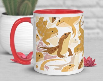 Puede incluir: Una taza de cerámica blanca con un borde rojo presenta un patrón de dragones barbudos de dibujos animados en varias poses. La taza está llena de un líquido marrón claro.