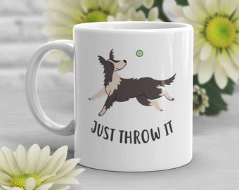 May include: White ceramic mug featuring a black and white illustration of a dog catching a green ball. The text "JUST THROW IT" is printed below the dog.