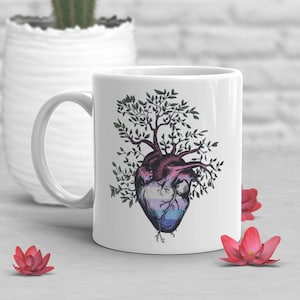 May include: White ceramic mug with a graphic of a heart with a tree growing out of it. The heart is colored in shades of pink, blue, and white. The tree has green leaves.
