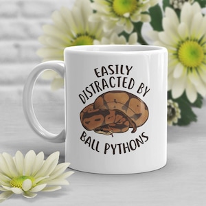 Op de afbeelding: Witte keramische mok met de tekst "EASILY DISTRACTED BY BALL PYTHONS" in het zwart. De mok heeft een bruine en bruine balpython-illustratie. De mok heeft een wit handvat en is omgeven door witte bloemen.