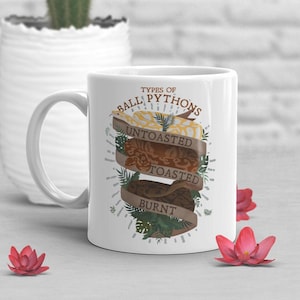 Könnte beinhalten: Weiße Keramik-Tasse mit einem Design von Königspythons, beschriftet mit "Untoasted", "Toasted" und "Burnt". Die Tasse hat einen Henkel und ist mit botanischen Elementen verziert. Der Text "TYPES OF BALL PYTHONS" befindet sich oben.