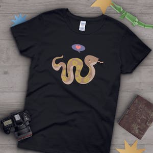 Op de afbeelding: Zwart T-shirt met een cartoon slang met een hart boven zijn hoofd. De slang is bruin en geel met een lichtblauwe gedachteballon boven zijn hoofd.