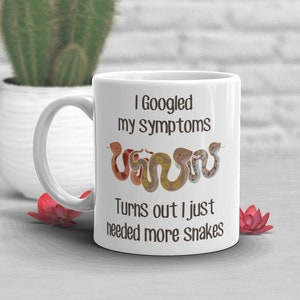 Könnte beinhalten: Weiße Keramik-Kaffeetasse mit braunem Textdesign, das "Ich habe meine Symptome gegoogelt Es stellte sich heraus, dass ich einfach mehr Schlangen brauchte" lautet. Die Tasse hat ein Cartoon-Bild von vier Schlangen in verschiedenen Farben und Mustern.