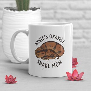 May include: White ceramic mug with a brown and tan snake illustration and the text "World's Okayest Snake Mom".