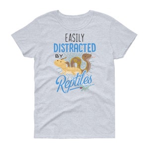 Może przedstawiać: Jasnoszary t-shirt z napisem "Easily Distracted By Reptiles" w kolorze niebieskim. Tekst jest otoczony wzorem promieni słonecznych. Są ilustracje węża i jaszczurki.