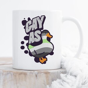 May include: White ceramic mug with a black and white illustration of a duck with green, gray, and black stripes. The text "gay as" is written in black above the duck.