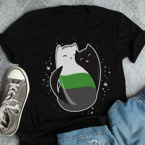 Könnte beinhalten: Schwarzes T-Shirt mit einem weißen und schwarzen Katzendesign. Die Katzen umarmen sich und haben ein grünes, graues und weißes Streifenmuster auf ihren Körpern.