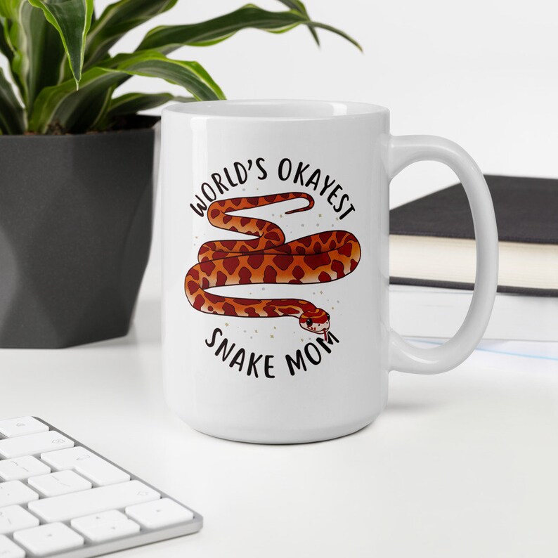 May include: White ceramic mug with a black and white graphic of a snake with the text "World's Okayest Snake Mom".