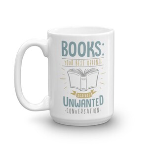 Puede incluir: Taza de cer&aacute;mica blanca con un asa grande. La taza presenta el texto "BOOKS: YOUR BEST DEFENSE AGAINST UNWANTED CONVERSATION" en letras azules y doradas, con una ilustraci&oacute;n de un libro abierto.