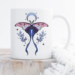 Peut inclure: Mug en céramique blanche avec un motif de papillon de nuit coloré. Le papillon a des ailes roses, violettes et bleues, un croissant de lune et des étoiles. Des brins de feuilles bleues et des étoiles ornent le motif. La tasse a une anse incurvée.