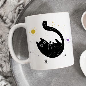 Op de afbeelding: Witte keramische mok met een zwart kattenontwerp en een sterrenhemelpatroon. De kat is wit omlijnd, met sterrenbeelden en hemelse elementen in geel en paars. De mok heeft een gebogen handvat.