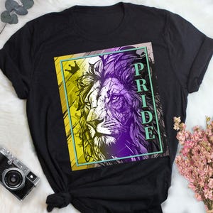 Puede incluir: Camiseta negra con un gráfico de una cabeza de león con un esquema de color amarillo y morado. La palabra "PRIDE" está escrita verticalmente en el lado derecho del gráfico.