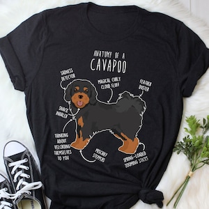 May include: A black t-shirt with a white graphic of a Cavapoo dog. The graphic includes text that says "Anatomy of a Cavapoo" and labels different parts of the dog, such as "Magical Curly Cloud Fluff", "Feather Duster", "Snack Inhaler", "Thinking About Velcroing Themselves to You", "Mischief Steppers", and "Spring-Loaded Zooming Sticks".