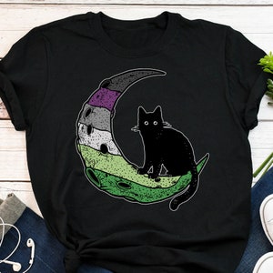 May include: Black t-shirt featuring a green, white and purple crescent moon with a black cat sitting on top. The moon has a textured design.