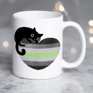 Puede incluir: Taza de cerámica blanca con un gato negro sobre un diseño en forma de corazón. El corazón presenta un patrón de rayas en tonos grises, negro y una franja verde. La taza tiene un asa curva.