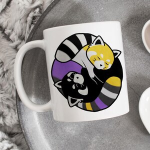 May include: White ceramic mug featuring a cartoon design of two red pandas in a yin-yang style. The design incorporates the colors black, yellow, purple, and gray. The mug has a curved handle and is set on a gray surface.
