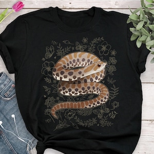 Peut inclure: T-shirt noir avec une illustration détaillée d'un serpent enroulé. Le serpent présente des marques marron et beige, entouré de motifs floraux et de papillons. Un t-shirt graphique élégant.
