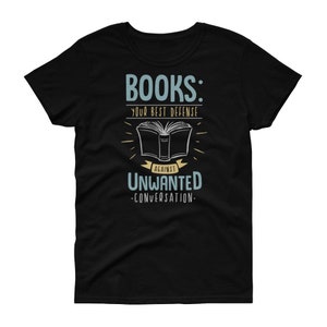 以下が含まれることがあります： 黒いTシャツに、白いグラフィックデザインが施されています。「本：望まない会話に対する最高の防御」と書かれています。グラフィックには、スタイリッシュな本の画像も含まれています。