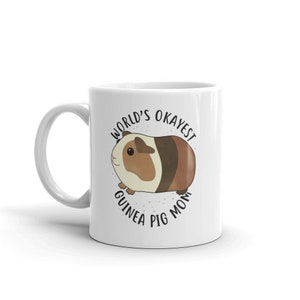 May include: White ceramic mug with a brown and white guinea pig illustration. The text "World's Okayest Guinea Pig Mom" is printed in black around the guinea pig.