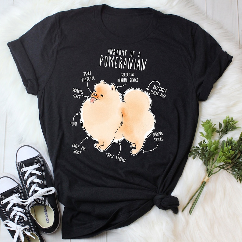 K&ouml;nnte beinhalten: Ein schwarzes T-Shirt mit einer wei&szlig;en Illustration eines Zwergspitz-Hundes. Die Illustration ist mit humorvollen anatomischen Merkmalen beschriftet, wie z. B. "Leckerli-Detektor", "T&uuml;rklingel-Alarm", "Obsz&ouml;n flauschiger Bereich" und "Snack-Lagerung". Der Text "Anatomie eines Zwergspitzes" befindet sich oben auf der Illustration.