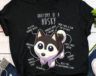 Puede incluir: Una camiseta negra con un gráfico blanco de un husky de dibujos animados. El gráfico incluye texto que dice "Anatomía de un Husky" y enumera varias características del husky, como "dispositivo auditivo selectivo", "pensando en travesuras", "pelusa mágica infinita" y "barriga llena de artículos cuestionables".