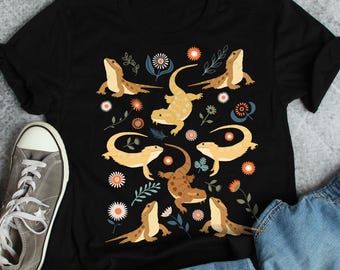 May include: Black t-shirt with a colorful pattern of bearded dragons and flowers. The bearded dragons are in various poses, some are facing forward, some are facing sideways, and some are facing backward. The flowers are in a variety of colors, including pink, yellow, and blue.