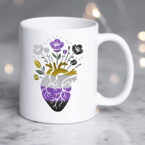 May include: A white ceramic mug featuring a graphic illustration of an anatomical heart. The heart is colored with horizontal stripes of yellow, grey, white, purple, and black. A bouquet of flowers in matching colors emerges from the top of the heart.