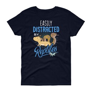 Może przedstawiać: Kobieta t-shirt w kolorze granatowym z napisem "Easily Distracted By Reptiles" w białym i niebieskim kolorze. Na koszulce znajduje się kresk&oacute;wkowa ilustracja węża, jaszczurki i gekona.