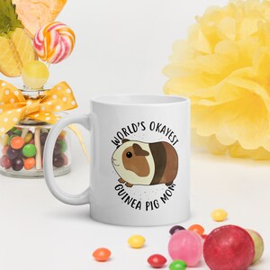May include: White ceramic mug with a brown and white guinea pig illustration and the text "World's Okayest Guinea Pig Mom".