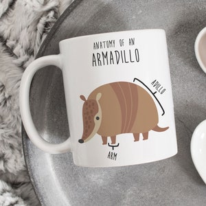 May include: White ceramic mug with a brown cartoon armadillo illustration. The mug has black text that reads "Anatomy of an Armadillo" and labels the armadillo's body parts "AdILLO" and "ARM".