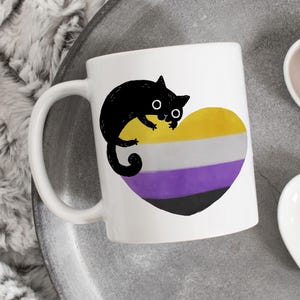 May include: White ceramic mug featuring a black cat illustration over a heart-shaped design in yellow, white, purple, and black, representing the non-binary flag. The mug has a curved handle and is set on a gray surface.