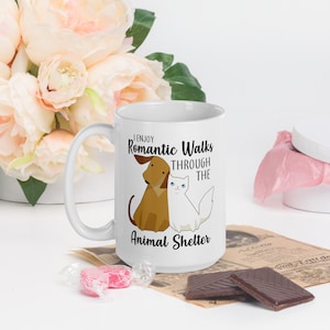 May include: White ceramic mug with a black and brown illustration of a dog and cat. The text on the mug reads "I enjoy romantic walks through the animal shelter."